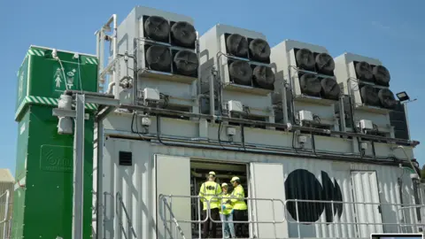 Jamie Niblock/BBC The Direct Air Capture plant seen from the outside, built around a grey shipping container. Three people in high-vis outfits stand by the open double doors. On top of the container are four large units each containing four black circular fans. To the left is a tall green rectangular unit with health and safety symbols on.