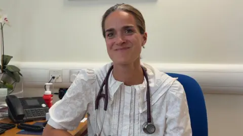 Dr Neele Hamilton sits in an office wearing a stethoscope. She has tied-up brown hair and a white blouse. She is smiling at the camera.