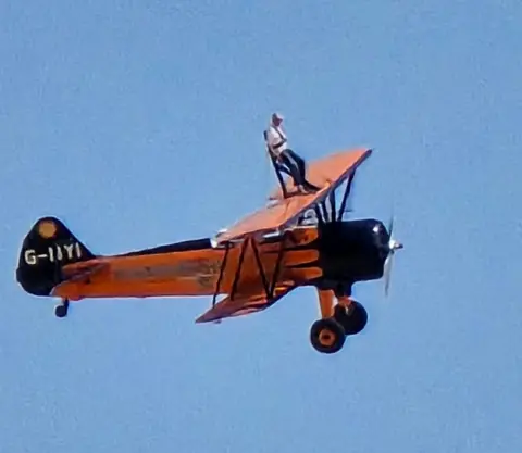 Clive England A man in a white t-shirt and blue trousers stands on top of an orange bi-plane in flight. He is harnessed to a frame. There is blue sky in the background.