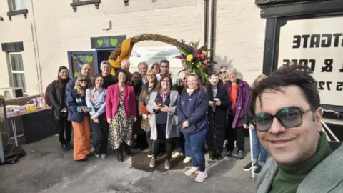 A group photo of people who attended the launch of the city fridge surplus food initiative at the Westgate Hotel in Oxford. Founder Saman Jamshidifard is taking the picture and smiling for the camera. Green hearts, which are part of the initiative's branding, can be seen above the door.