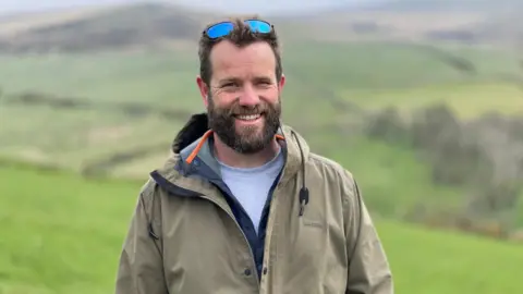 Carl Rowlinson has short brown hair and a beard. He is smiling standing in a field with rolling hills behind him. He has sunglasses on his head and is wering a khaki jacket.