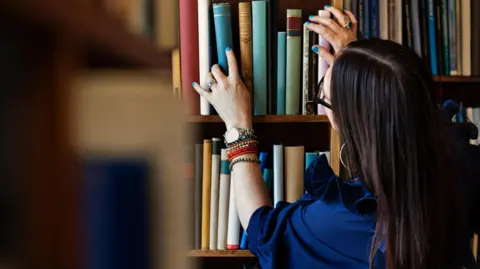 A stock image of a library worker, looking at books. She has long dark hair, is wearing a blue top, and has on a watch and bracelets on her left arm. She has a ring on her right hand, which is raised and on a book. The are a large number of books on shelves.