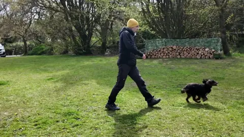 Roy Young walking across a field with a black cocker spaniel running in front.