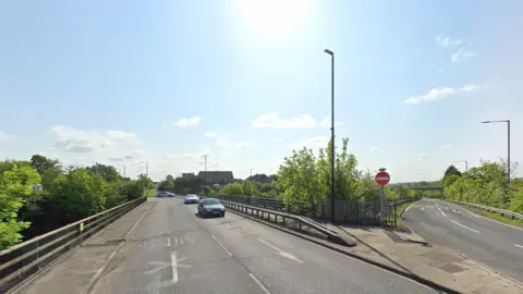 A two-lane road over the M1. It is a sunny day with blue skies. 