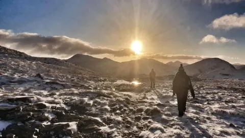 Steven Williamson Two hikers walk across a snow-dusted mountain trail as the sun rises over distant peaks, casting long shadows across the rugged terrain.