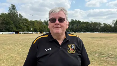 There is a man in a black polo shirt with yellow lines on the shoulders. He has two buttons undone. To the left of his shirt, it says the manufacturer VX3 and the right, the club's crest. He is wearing dark sunglasses and has short grey hair. Behind him, there is a pitch covered in brown grass. There is a rugby goal to his right and a floodlight to his left. 