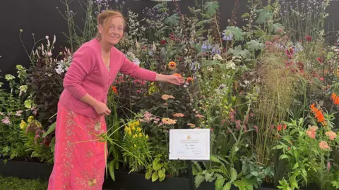 Michaela Boggis wears a pink cardigan a long jewelled pink skirt and turquoise crocks. She stands in front of a beautiful show garden of flowers featuring dahlias, agapanthus, berkheyas and crocosmia.