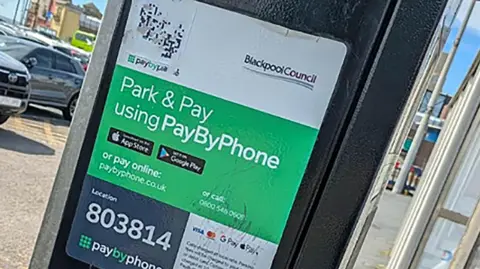 Local Democracy Reporting Service Close up of a car park payment machine in Blackpool's Central Car Park. Parked cars are in the background along with Coral Island arcade in the distance. It is a sunny day.