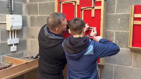 BBC/Molly Brewer An older man wearing a navy fleece and shirt, has his back to the camera as he works on a construction board with pipes. Next to him is a young girl, with short brown hair, wearing a blue coat, also working on a piece of wood and pipe with the teacher.