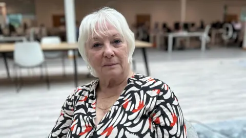 Kate Bradbrook/BBC Danielle Stone in a black, white and red patterned blouse looks directly at the camera. The background of the image is in soft focus placing emphasis on Ms Stone.