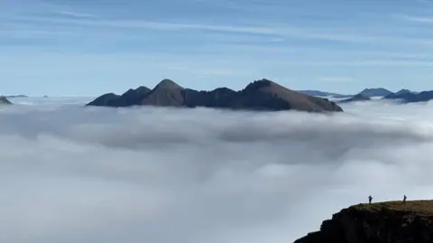 Willie Baird Jagged mountain tops rise above a thick blanket of clouds under a clear blue sky.