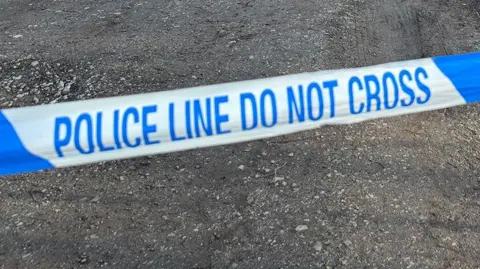 Blue and white police tape used to cordon off a crime scene. The ground is muddy with gravel.