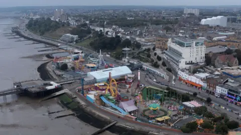 Getty Images A drone aerial view of Southend-on-Sea. The Adventure Island theme park is visible, with its red and yellow-painted rollercoaster ride and funfair attractions. The sea is to the left of the frame, and buildings are on the right. It is a cloudy day.