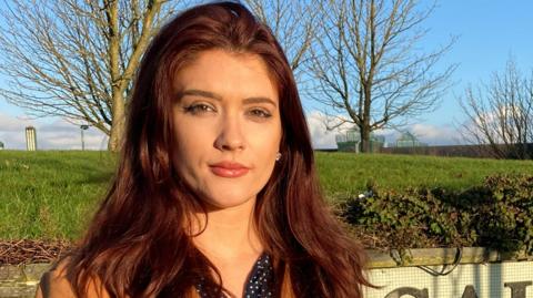 Cara Hunter, a woman with long reddish hair, outside with trees in the background. She is looking directly at the camera and is not smiling.