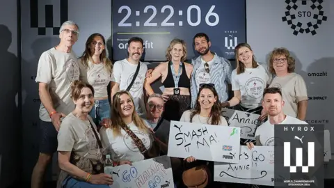 Hyrox/Annie Reid A group of people wearing matching white T-shirts and holding signs smile at the camera. Some of the group are kneeling at the front. There is one woman in the middle wearing a black crop top and leggings with a medal around her neck. Above them is an electronic sign displaying the time 2:22:06.