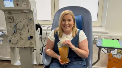 Family Handout/ NHS Blood and Transplant/PA Wire A woman with blonde hair a white tshirt and blue top in a blue chair holding a plastic bag containing a pale brown liquid