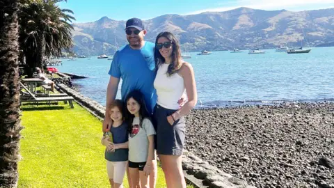 A man, a woman and two young girls standing next to a lake with mountains in the background. It is sunny - the adults are wearing sunsglasses. 
