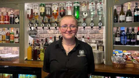 BBC/ Alex James A woman with short blonde hair is stood behind a bar wearing a black shirt with a pub logo on the front. She is stood in front of a number of bottles of alcohol.