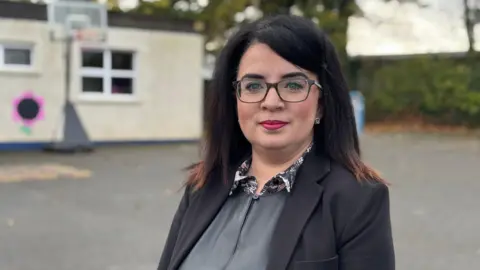 A woman stands in the playground of a primary school. She has dark hair and glasses and has bright red lipstick on. She's smartly dressed in a grey shirt and a black blazer. She has a polite smile. 