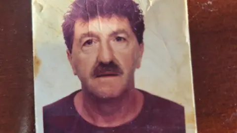 A passport-style creased photograph of a man with dark hair and a moustache. He is staring into the camera. 