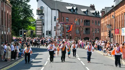 Pacemaker A wide shot of the parade in Ballymena, Orangemen are walking along the road with a banner while people stand on either side of the road to watch. There are terraced red brick buildings on each side of the road and a more modern block of flats behind.