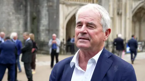 Allan Lamb, wearing a blue jacket and white shirt standing outside Winchester Cathedral