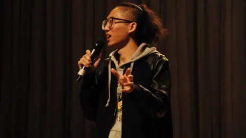 Supplied Chizi performs on stage in 2016, wearing a grey hoodie under a black jacket, with his long hair tied back.