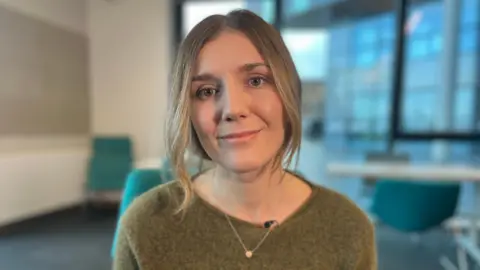 A head and shoulders photograph of Dr Amber Phillips. She is wearing a light brown jumper and has shoulder-length light brown hair. She is sitting in a room with a white table and green chairs behind her