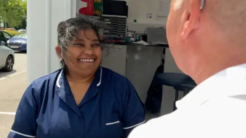 Irene is talking to someone - only the back of their head is visible in the foreground. She is wearing a blue lab coat. Behind her the interior of the testing van can be seen with papers and cabinets.