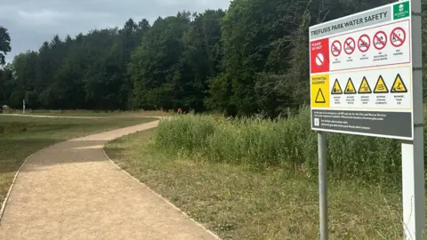 A picture taken from the entrance to Trefusis Park. There is a big sign advising water safety and a large path carved around grass with large green trees in the background.