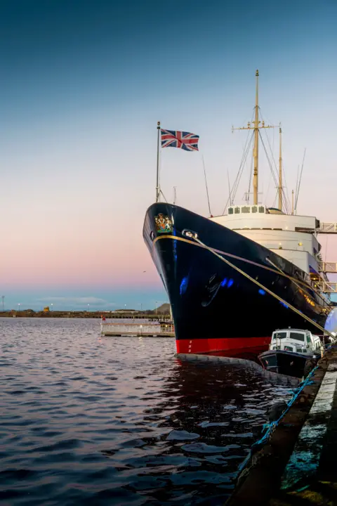 Tony Marsh A large boat sits in the water with the sun setting behind it. The boat is red, black and white and has a Union Jack flying on a flag pole on the front.