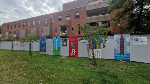 The artworks on grey hoardings in front of the building featuring Grimsby Minster, a wind turbine, Grimsby Town's badge a rainbow and the Dock Tower