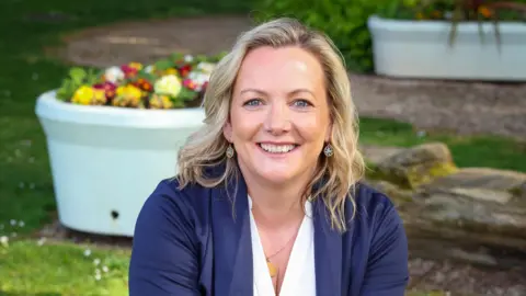 Mary McFarland looking to the camera and smiling.  She has shoulder-length, wavy blonde hair and blue eyes.  She is wearing a navy cardigan over a white v-neck top. 