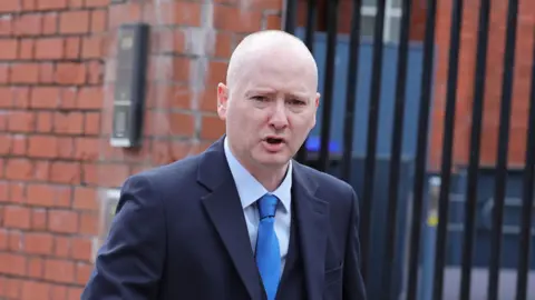PA Media A bald headed man in a dark suit with a blue shirt and tie