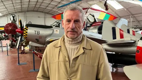 Shaun Whitmore/BBC Paul Hennessey, wearing a polo neck jumper and tan winter coat, is looking at the camera. Behind him is a World War 2 plane and museum artefacts. 