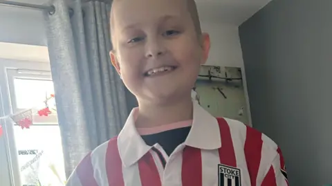 Family photo Harrison looks at the camera and smiles. He wears a red and white Stoke City football shirt over a black t-shirt with a pink collar. Behind it are curtains and window decorations. 