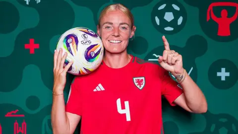 Getty Images Sophie Ingle of Wales poses for a portrait in her Wales shirt holding a football with her right hand. She has a left hand finger pointing up the sky. The background is a dark green colour.