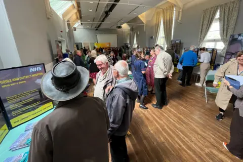 BBC/Anne-Marie Tasker People standing in a room at a drop-in session. There is a wooden floor and a display on a table with "NHS" written on it.