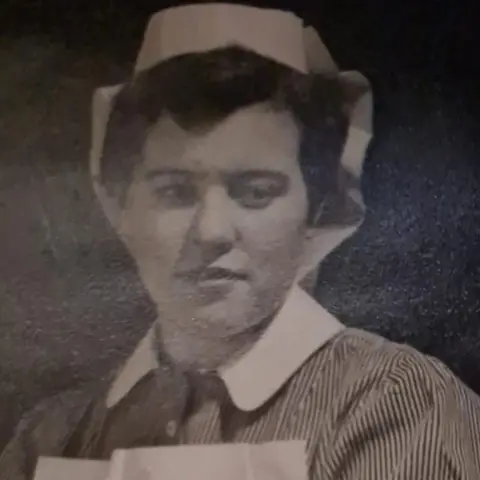 Fiona Brown A black and white photograph of an adult Moira wearing nurse scrubs.