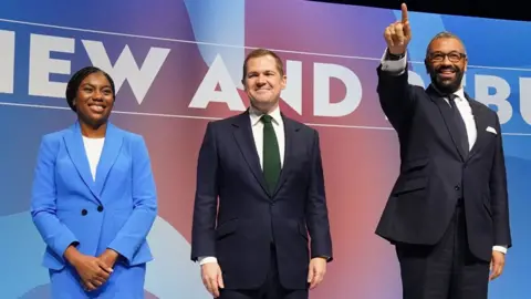 PA Media De twee overgebleven kandidaten, Kemi Badenoch in een felblauw pak, Robert Jenrick in een marineblauw pak en Cleverly in een marineblauw pak, stonden naast elkaar op het podium van de Conservatieve Partij-conferentie - allemaal glimlachend naar het publiek.