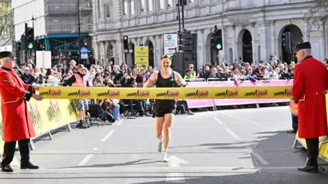 PA Media Runner Stefan Otway smiles as he crosses the finish line, breaking a yellow ribbon held by two officials in formal red coats.