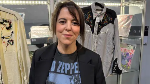 A woman with brown hair tied back. She is wearing a black jacket on top of a blue and black t-shirt which has Led Zeppelin written on it, which is partly visible. 