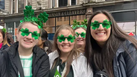 Lively group is gathered in front of a building with the sign 'ROBINSON & CLEAVER LTD.' The scene is vibrant, with people dressed in festive green attire and accessories like shamrock-themed headbands and glasses reflecting a celebration of St. Patrick's Day. In the background, the city street buzzes with activity, featuring other buildings and pedestrians.