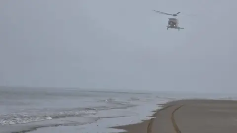 Wirral Coastguard A helicopter hovers over the shoreline on the beach.
