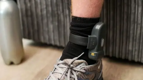 BBC/Rare TV Image of someone's lower leg with an ankle tag above the sock.