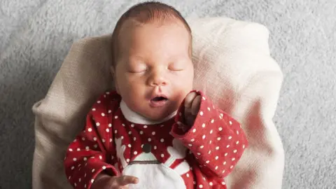 A baby wearing a Christmas jumper lying on a white blanket, with its left hand raised towards it mouth 