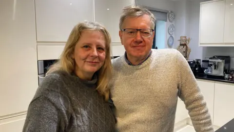 A woman with long blonde hair wearing a dark grey sweater stands in a kitchen with her arm around a man with short grey hair and glasses. He is wearing an off-white sweater.