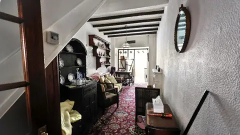 Paul Fosh Auctions A general viewof the lounge in the house. Wooden shelving, tables and chairs are on either side of the narrow room. At the far end of the room, there is a open partition into the kitchen. 