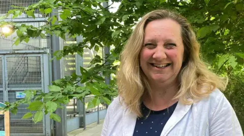 Lucy Nethsingha is wearing a white jacket with a blue and white dress. She has shoulder length blonde hair and is smiling at the camera