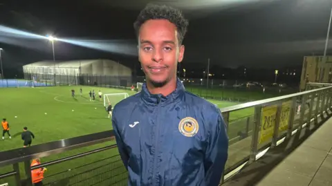 Warsome Hersi standing on a balcony at the football grounds in Lockleaze. He s wearing a dark-blue sports jacket. There are a group of men playing football on the pitch in the background on a dark evening.